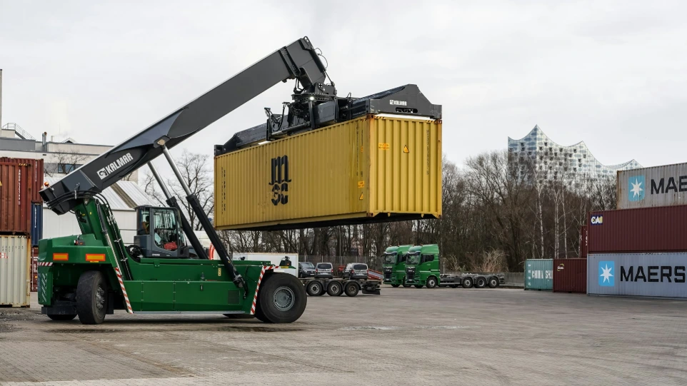 A green reach stacker lifting a yellow shipping container.